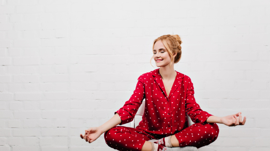 Pleased girl in red pajamas doing yoga on white background. Indoor portrait of blonde young lady sitting in lotus pose on chair.